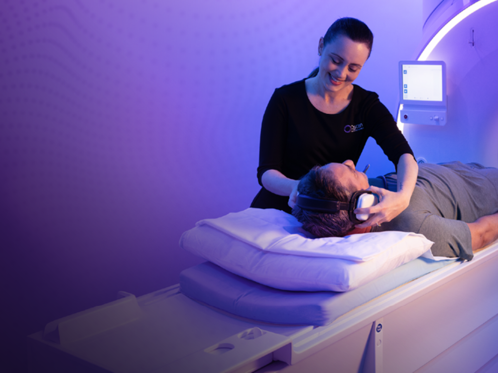 A smiling Qscan staff member places headphones on a patient lying on an MRI scanner table, ready for their scan, in a room lit with soft purple light.