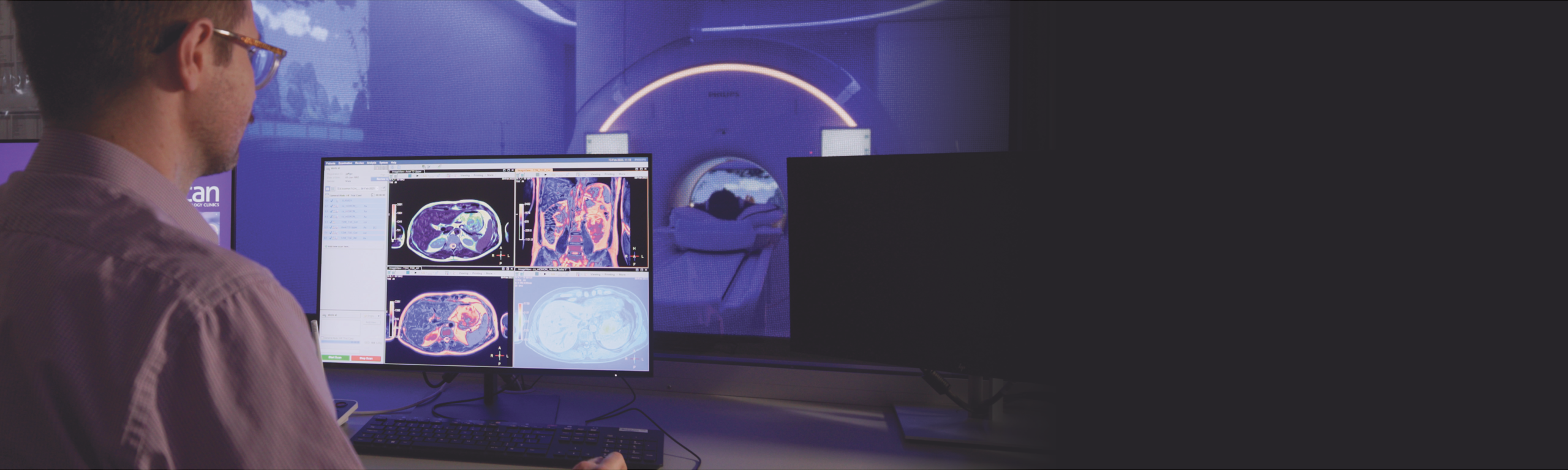 A radiographer reviews a patient scan, while a patient has an MRI in the background
