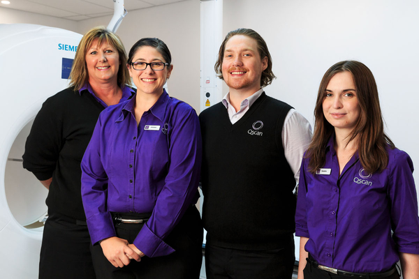 "Four smiling Qscan staff members in purple and black uniforms standing beside a CT scanner at the Qscan North Hobart clinic.