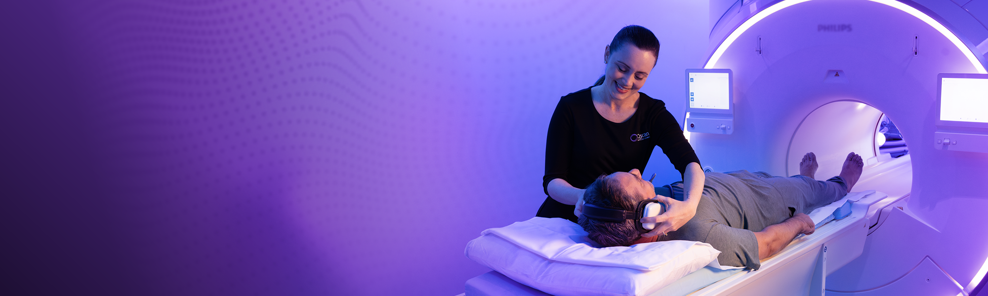 A smiling Qscan staff member places headphones on a patient lying on an MRI scanner table, ready for their scan, in a room lit with soft purple light.